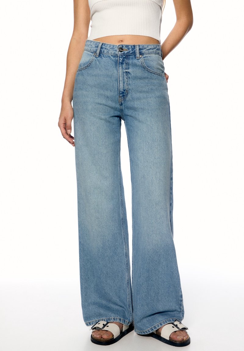 Femme portant un jean taille haute à jambes larges bleu clair, un haut blanc court sans manches et des sandales blanches avec des semelles noires.