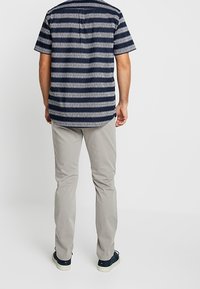 Tmavě modrá košile s krátkými rukávy s bílým proužkovaným vzorem, doplněná světle šedými kalhotami a tmavě modrými teniskami, pohled zezadu.