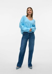 Hellblauer, lockerer Strickpullover mit V-Ausschnitt und Rautenmuster, kombiniert mit mittelblauen Jeans im geraden Schnitt und blauen High Heels.