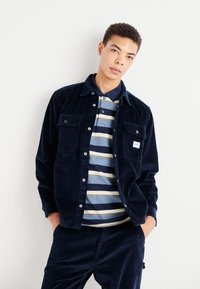 Giacca camicia in corduroy blu navy con due tasche sul petto, indossata sopra un polo a righe in blu, navy e beige. Vestibilità slim con una consistenza morbida.