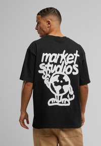 Sort bomulds T-shirt med stort hvidt tekst "market studios" og en tegneserie-verden karakter på ryggen. Korte ærmer, afslappet pasform.
