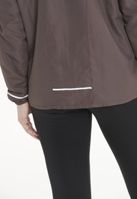 Brun lätt jacka med en jämn textur, med en vit accent vid fållen och ärmen. Bärs över svarta leggings.