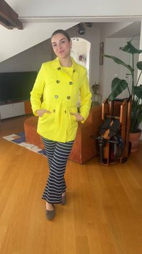 Femme souriante à l'intérieur portant un manteau jaune vif et un pantalon à motifs, debout sur un sol en bois près d'une plante et d'un appareil d'exercice plié.