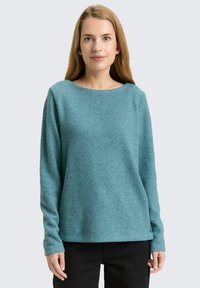 Vrouw met lang blond haar, gekleed in een teal gebreid sweater en zwarte broek, staande tegen een effen lichtgrijze achtergrond.