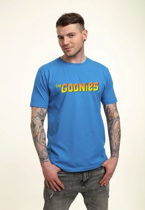 Mand med kort hår og tatoveringer på begge arme bærer en lyseblå t-shirt med "THE GOONIES" i fede gule og orange bogstaver.