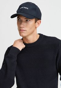 Svart caps med "Casual" trykt i hvit tekst, kombinert med en teksturert svart genser med et sikk-sakk mønster. Uformelt design, tettsittende fasong.