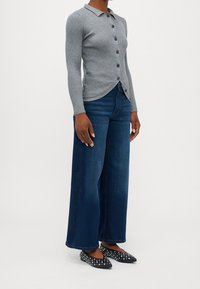 Žena v šedém žebrovaném cardigan s knoflíky, v širokých tmavě modrých džínách a černých bytelových jehlových balerínách, stojící na jednobarevném pozadí.