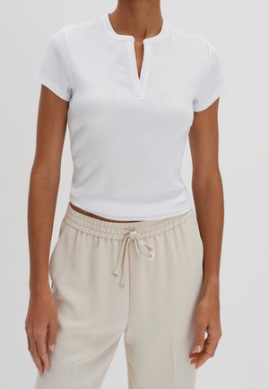 Vrouw draagt een wit, geribbeld kortemouwtopje met een V-hals en beige broek met trekkoord, staand tegen een effen achtergrond.