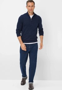 Marineblauer Pullover mit Reißverschlusskragen, langen Ärmeln und gerippten Bündchen, über einem weißen Hemd getragen, kombiniert mit dunklen Jeans und braunen Stiefeln.