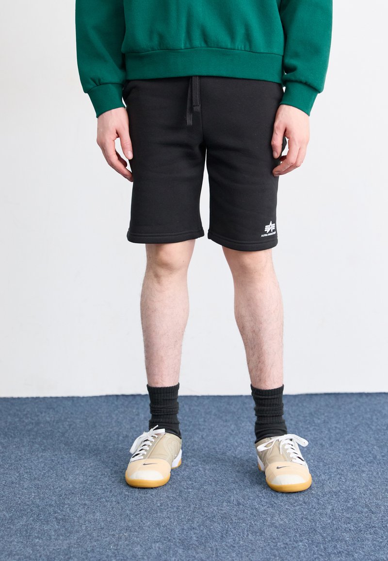 Shorts de maillot noir avec une ceinture élastique, ornés d'un logo sur l'ourlet inférieur. Associés à des baskets beiges, blanches et jaunes et des chaussettes noires.