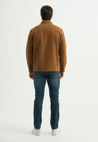 Veste décontractée marron, à manches longues, texture lisse, coupe slim. Portée avec un jean bleu et des baskets blanches, avec des détails de couture visibles.
