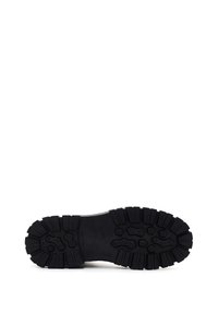 Suela de un zapato negro con surcos profundos y patrones rectangulares y hexagonales para mejorar el agarre, fotografiada sobre un fondo blanco.