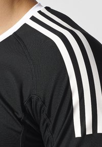 Maillot de sport noir en tissu texturé, avec un design à trois rayures blanches sur les épaules et un col rond.