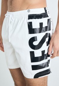 Shorts de bain blancs avec un grand logo noir imprimé sur le côté. Le matériau est lisse, avec une taille élastique et un cordon de serrage.
