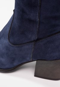 Mörkblå svinskon ankle boot med klack och synliga sömmar på sidan, vilande på en vit yta.