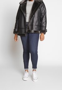 Persona que lleva una chaqueta de shearling negra sobre un suéter de punto gris, jeans skinny oscuros arremangados y zapatillas blancas con cordones.
