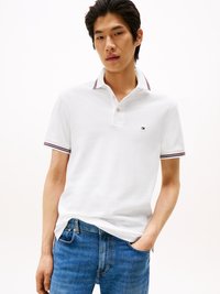 Junger Mann in einem weißen Poloshirt mit gestreiftem Kragen und B�ündchen, kombiniert mit blauen Jeans, steht mit einer Hand in der Tasche.