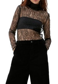 Schwarzes Spitzenoberteil mit langen Ärmeln, das über einem soliden schwarzen Panel auf der Brust verfügt. Strukturiertes Blumenmuster, hoch geschnittenes Design, halbtransparentes Material.