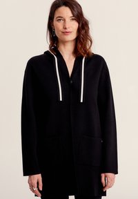 Cappotto lungo nero con cappuccio e due tasche frontali. Presenta cordini a strisce bianche, una chiusura con bottoni e una vestibilità rilassata. Texture morbida.
