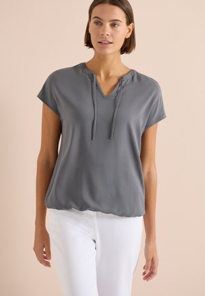 Kobieta ubrana w szarą bluzkę z krótkim rękawem, dekoltem w serek i sznureczkami, w połączeniu z białymi spodniami, stojąca na beżowym tle.