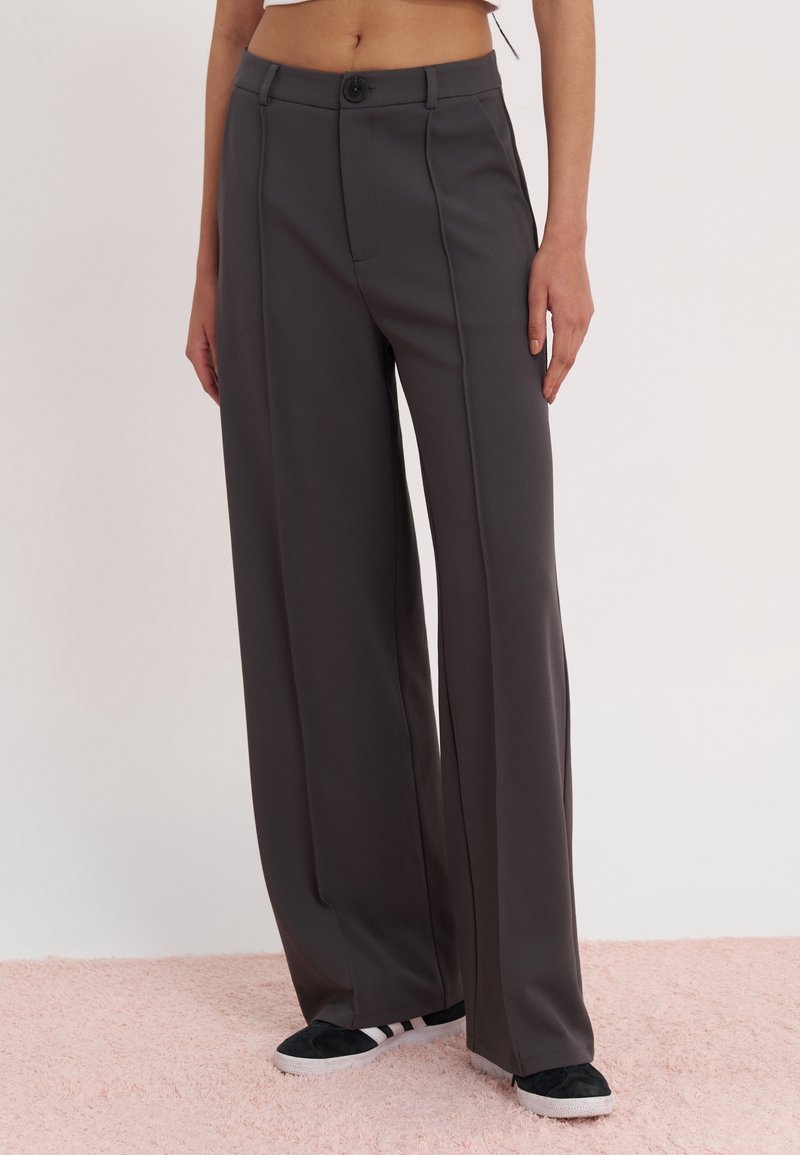Femme portant un pantalon large taille haute gris foncé et des baskets noires et blanches, debout sur un tapis rose clair.