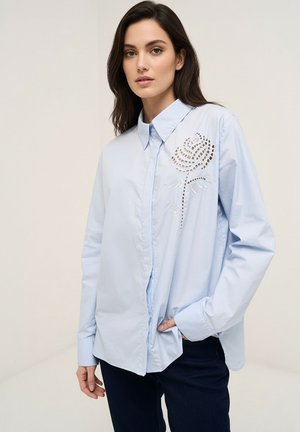 Kvinde iført lyseblå knap-op skjorte med blomstret udskæring og broderi, parret med mørke bukser, stående mod en ensfarvet baggrund.