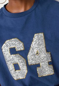 Camiseta azul marino con grandes números "64" en lentejuelas plateadas metálicas bordeadas en oro. Tejido suave, con manga corta y escote redondo.