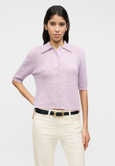 Ανοιχτό μωβ πλεκτό πόλο μπλουζάκι με κοντό μανίκι, με υφασμένο σχέδιο, πατιλέτα με κουμπιά και διπλωμένο γιακά, συνδυασμένο με κρεμ παντελόνια και μαύρη ζώνη.