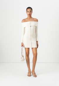 Off-shoulder ribbet strikket kjole i kremfarge, med frontglidelås og lange ermer. Kombinert med en liten håndveske og sandaler med stropper.