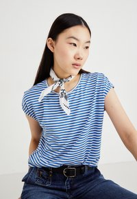 T-shirt rayé bleu et blanc avec des manches retroussées, associé à une ceinture noire et une écharpe blanche à motifs nouée autour du cou.