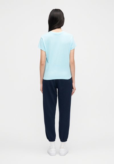 Gaiši zils īsroku t-krekls ar brīvu piegriezumu, kopā ar tumši ziliem jostasvietas biksēm ar elastīgām aprocēm, valkāts ar baltām sporta kurpēm.