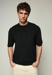 Sort strikket t-shirt med korte ærmer, ribbet rund hals og afslappet pasform, båret med hænderne i lommerne, sat mod en neutral baggrund.