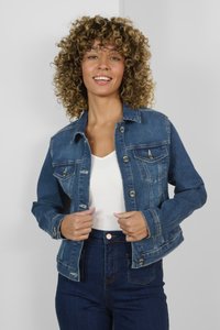 Chaqueta de mezclilla en azul medio, ligeramente desgastada, con un cierre de botones en la parte delantera, dos bolsillos en el pecho y un cuello estructurado. Lleva puesta sobre una blusa blanca.