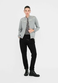 Giacca bomber grigia con perline decorative sulle spalle, abbinata a pantaloni jogger neri con tasche laterali e stivali neri texturizzati.