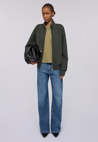 Grüne Reißverschluss-Bomberjacke, olivfarbenes Oberteil und weit geschnittene blaue Jeans, hält eine schwarze Tasche. Glatter Stoff, neutrale Farbpalette und lockere Passform.