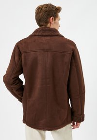 Chaqueta de ante sintético marrón con un cuello y puños de pelo texturizado, que presenta un corte recto y costuras en la parte posterior.