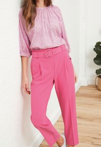 Blouse rose clair avec un motif texturé et un col froncé, associée à un pantalon rose vif avec une large ceinture et des poches latérales.