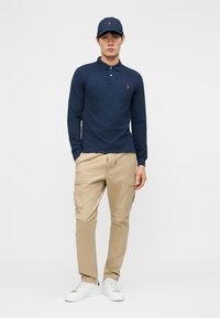 Vyras, vilkintis tamsiai mėlyną ilgomis rankovėmis polo marškinėlį, atitinkamą kepuraitę, smėlio spalvos cargo kelnes ir baltus sportinius batelius, stovintis su dalinai kišenėse įkištomis rankomis prieš baltą foną.