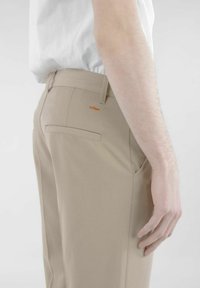 Beige Hosen mit slim Fit, ausgestattet mit einer einzelnen Gesäßtasche und einem orangefarbenen Markenlogo darüber. Glatte Stofftextur, minimales Design.