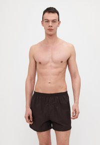 Shorts de bain en coton marron avec une taille élastique, un détail de bouton et une coupe ample, portés par un modèle se tenant devant un fond clair.