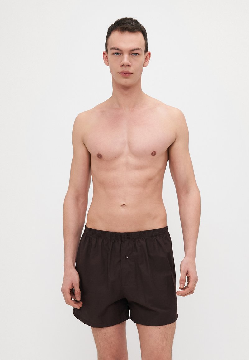 Shorts de bain en coton marron avec une taille élastique, un détail de bouton et une coupe ample, portés par un modèle se tenant devant un fond clair.
