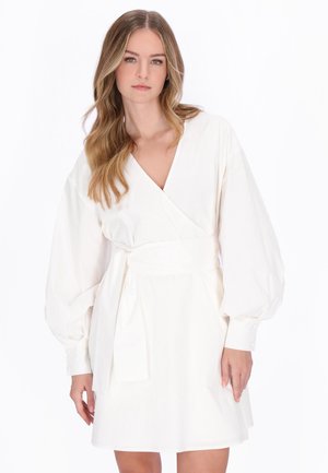 Młoda kobieta z długimi falującymi blond włosami, ubrana w białą sukienkę kopertową z długimi bufionymi rękawami na jednolitym białym tle.