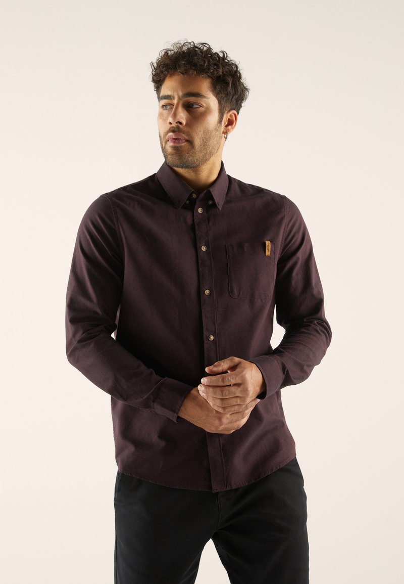 Dunkelviolette Button-up-Hemd, lange Ärmel, einzelne Brusttasche, lässiges Design. Texturierter Stoff mit braunen Knopfakzenten, in schwarze Hose gesteckt.