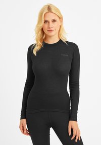 Young woman with blonde hair wearing a fitted black long-sleeve top and matching black pants, standing against a white background.