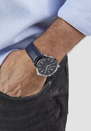 Analoge horloge met een zwarte, gestructureerde wijzerplaat, zilveren kast en blauwe leren band. Wordt gedragen om een pols, waarbij de hand in een zak rust.