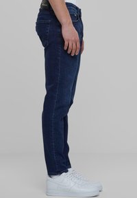 Sötétkék farmernadrág, slim fit szabású, öt zsebbel, varrott részletekkel és fekete címkével a derékpántnál. Fehér sportcipővel viselve.