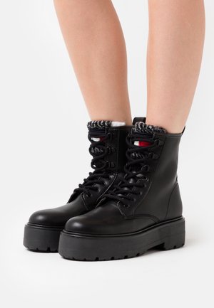Bottes à plateforme en cuir noir à lacets portées avec des chaussettes noires et blanches en tricot visibles au-dessus de la cheville sur des jambes nues.