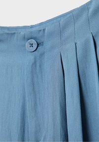 Gros plan sur un pantalon en tissu bleu clair montrant la taille, le bouton et les plis sur le côté droit.