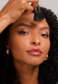 Femme aux cheveux noirs bouclés appliquant un fond de teint sur son front à l'aide d'un grand pinceau.