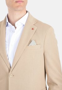 Blazer beige con trama, colletto a lancia, chiusura a bottoni e un'emblema decorativa rossa. Camicia bianca sotto con un fazzoletto a pois nella tasca.